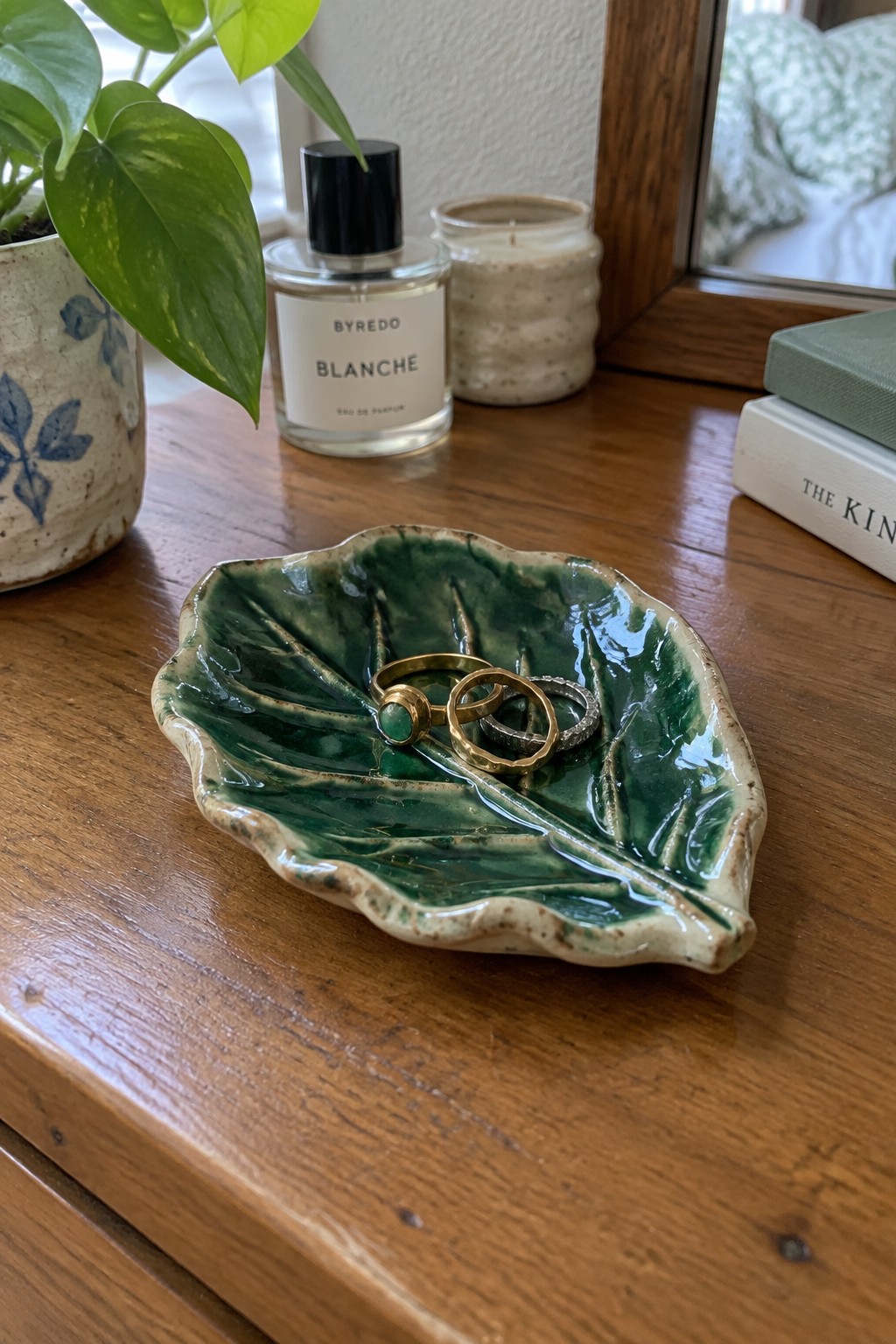 Green leaf-shaped ceramic dish holding rings beside Byredo Blanche perfume on wooden table