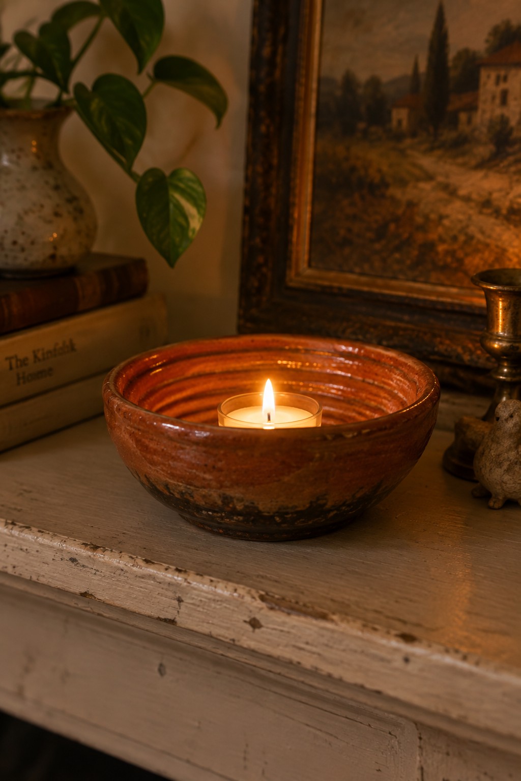 Lit candle in ribbed ceramic bowl on aged wood with books, plant, and painting nearby