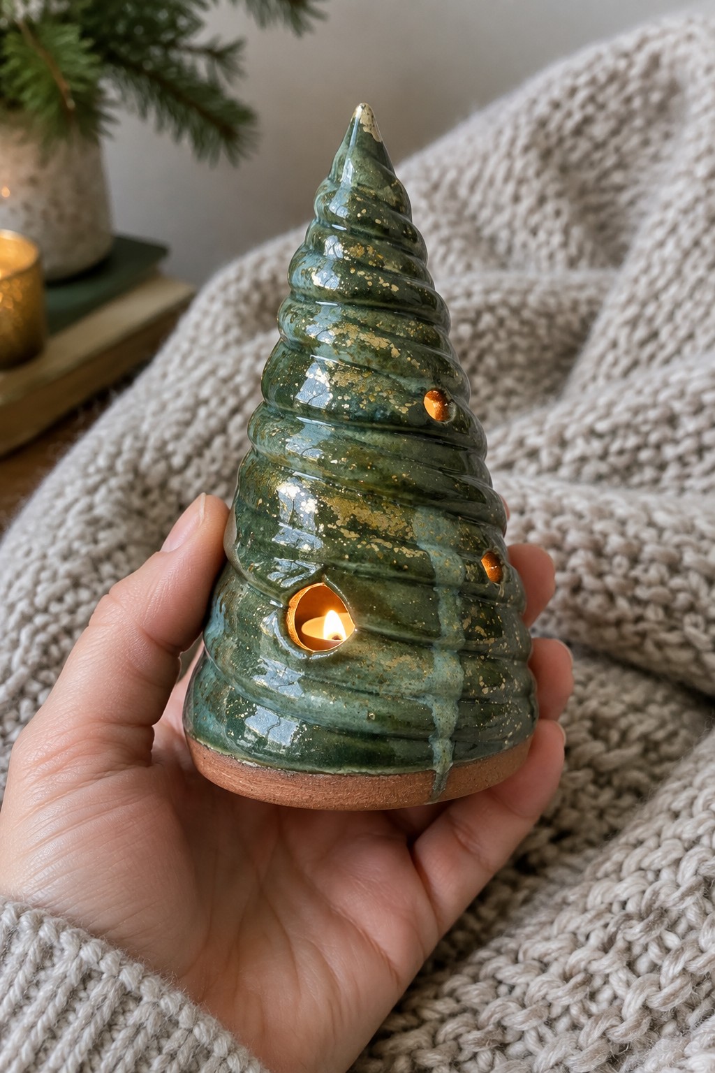 Hand holds green ceramic Christmas tree candle holder glowing with candle on beige knit blanket