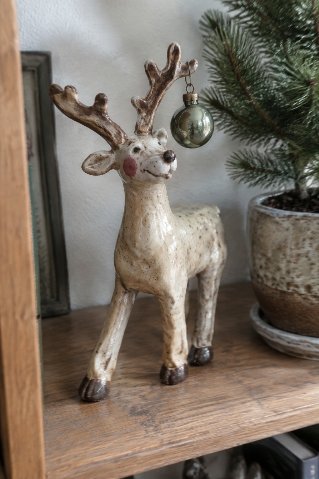 Ceramic reindeer figurine with speckles, pink cheek, and ornament on wooden shelf beside plant.