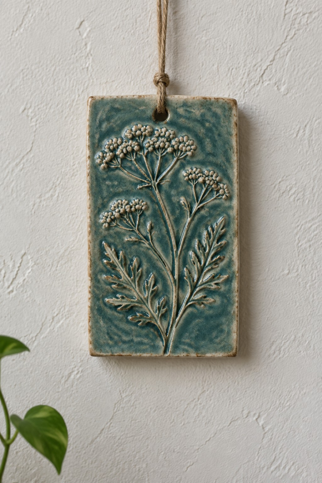 Teal ceramic tile with embossed plant design hanging by twine on white wall