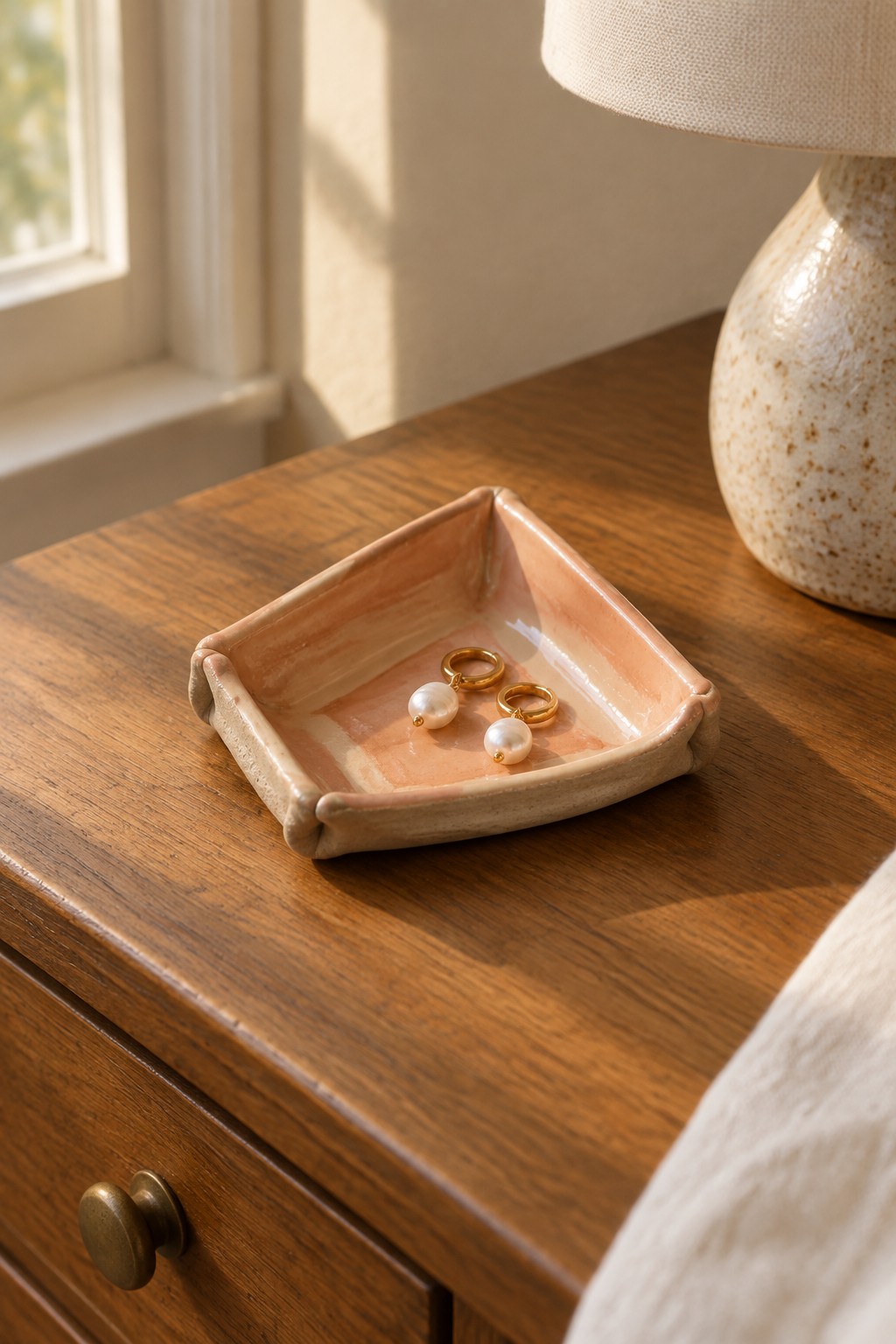 Peach ceramic tray holding pearl drop earrings on sunlit wooden nightstand
