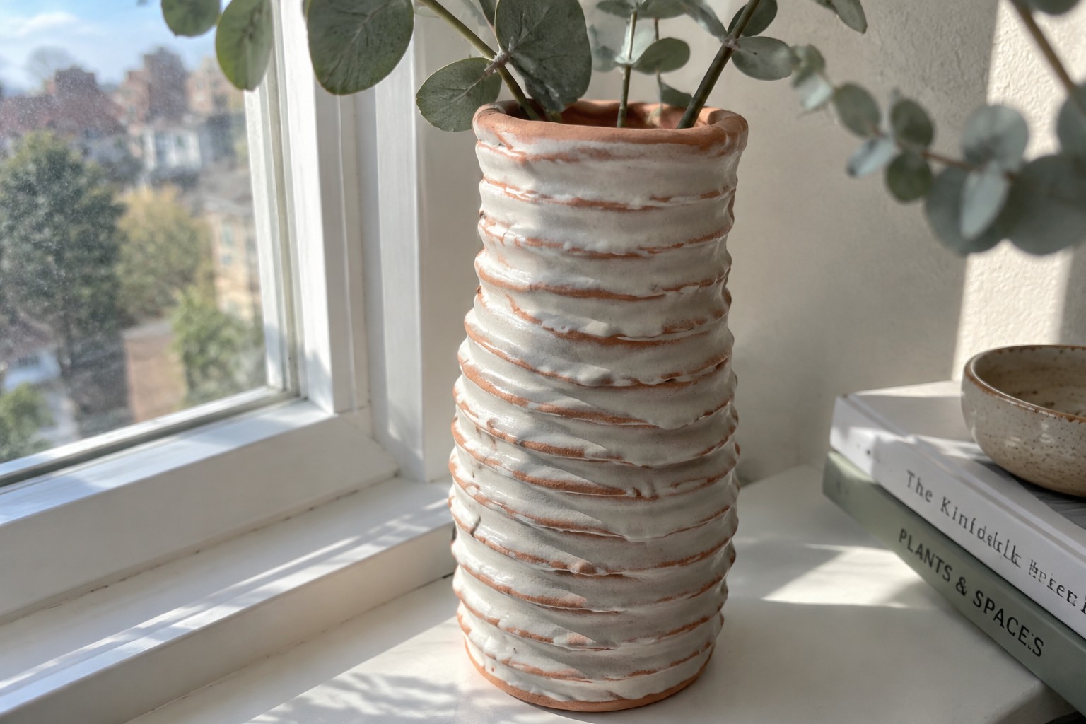 Tall white ribbed ceramic vase holding eucalyptus on sunlit windowsill with book