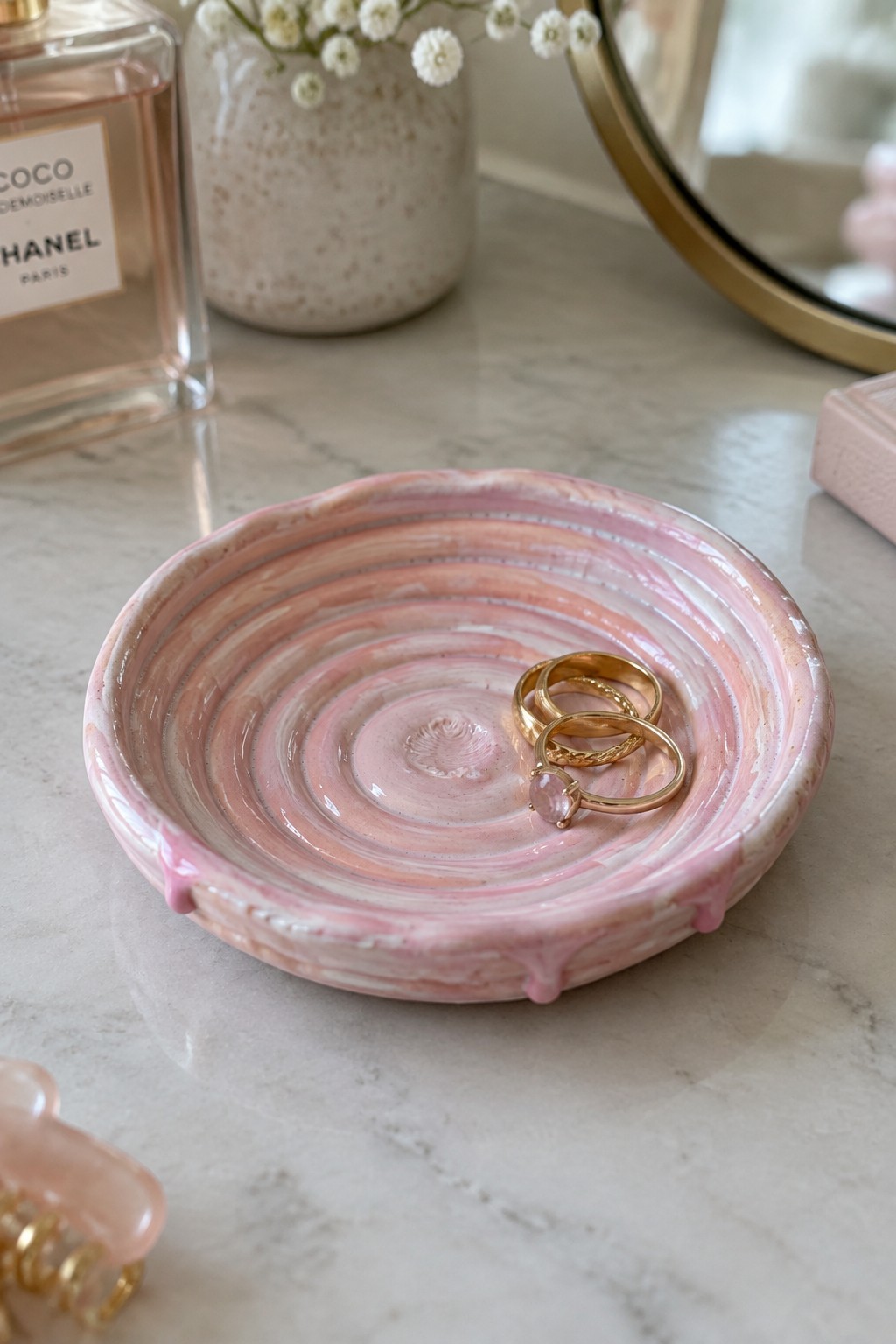 Pink swirled ceramic dish holding gold rings on marble vanity beside perfume
