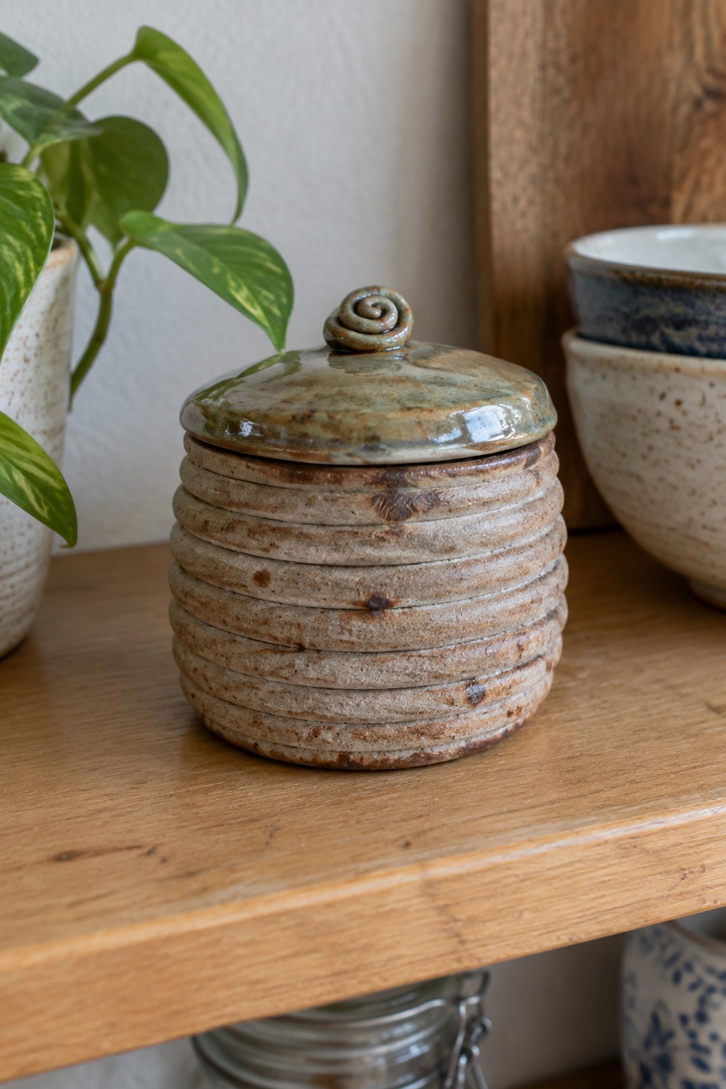 Rustic ribbed ceramic jar with spiral lid on wooden shelf beside plant and bowls