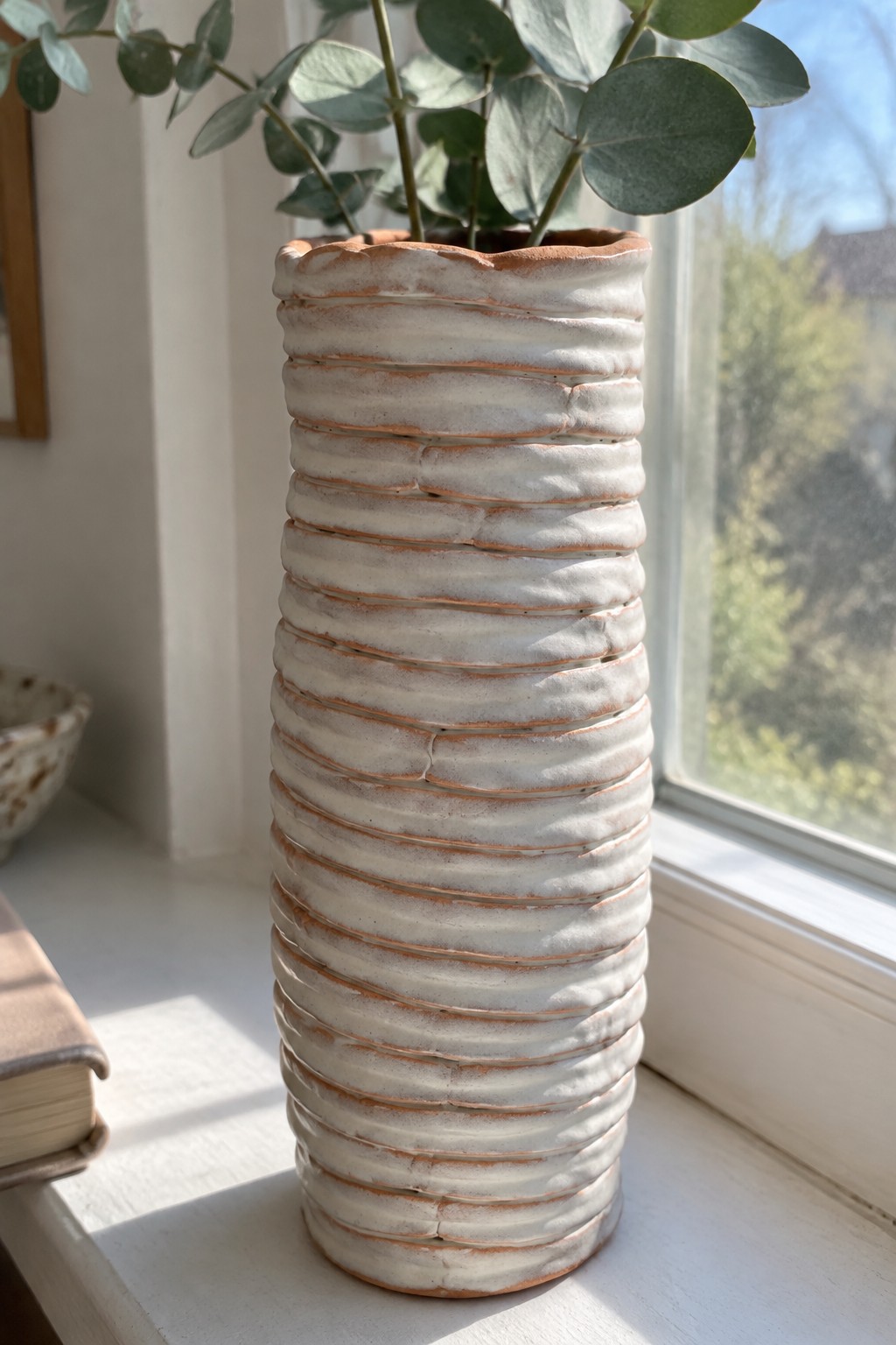 Tall white ribbed ceramic vase holding eucalyptus on sunlit windowsill with book