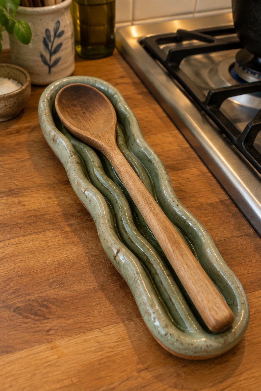 A wooden spoon rests inside a long, wavy green ceramic holder built from coiled clay on a wooden kitchen counter.