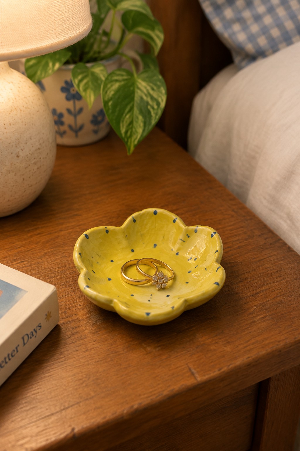 Yellow flower-shaped ceramic dish with blue speckles holding two gold rings on wooden nightstand.