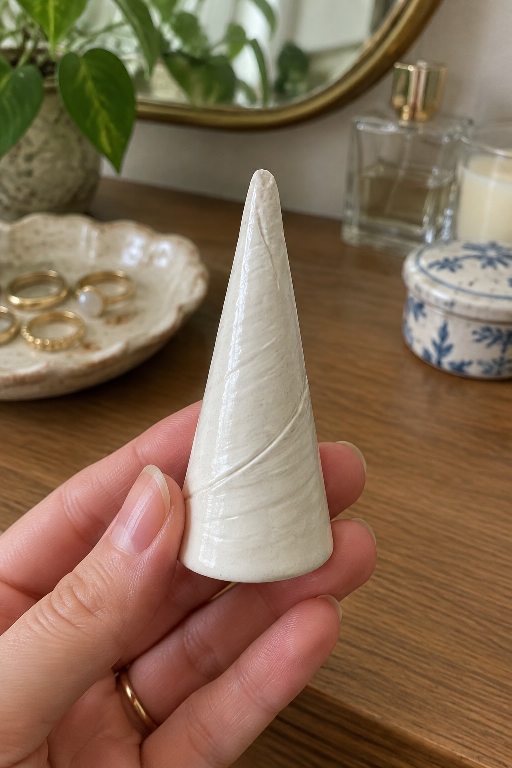 Hand holding glossy white ceramic cone before wooden vanity with jewelry and perfume