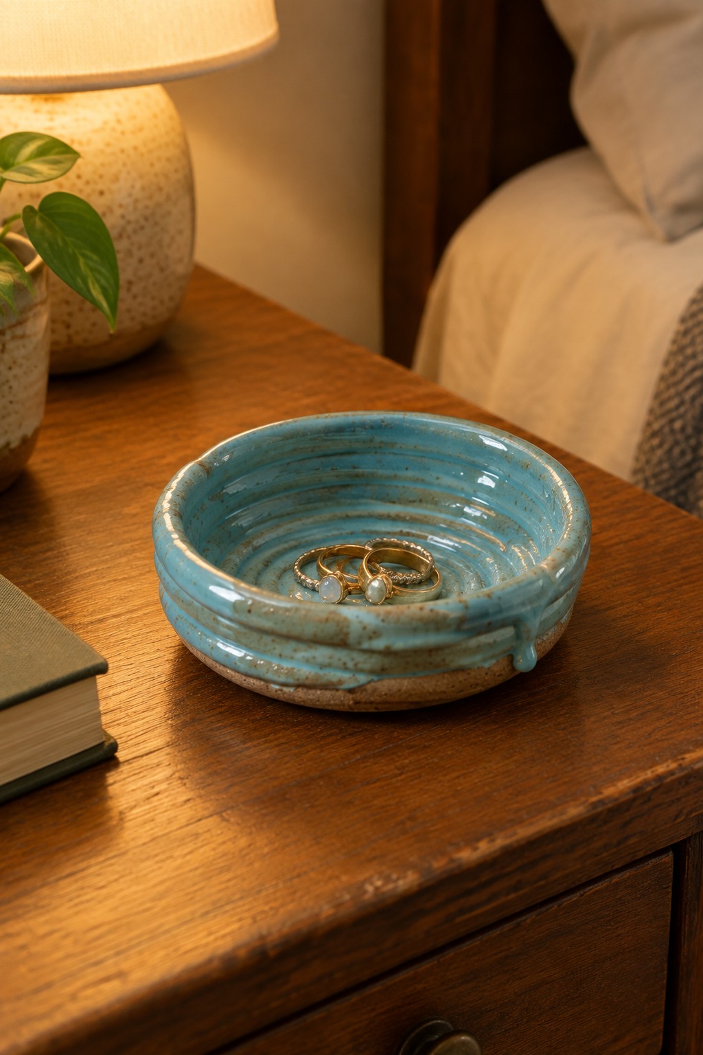 Turquoise ceramic bowl holding gold pearl rings on wooden nightstand beside lamp