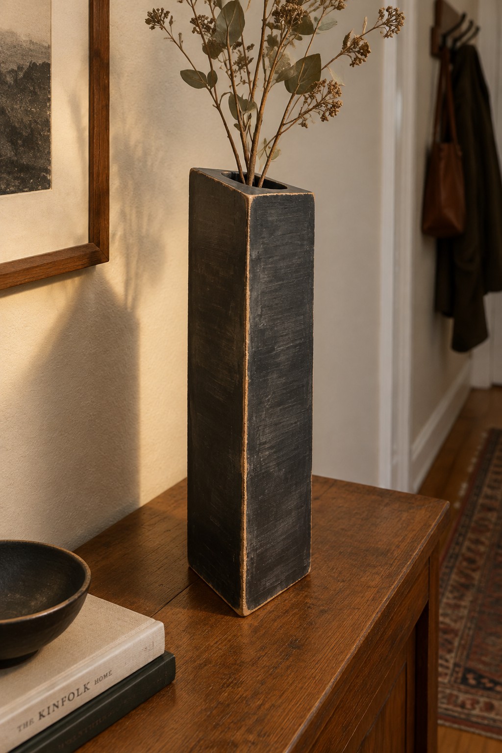Tall black vase with dried branches on wooden table beside bowl and books