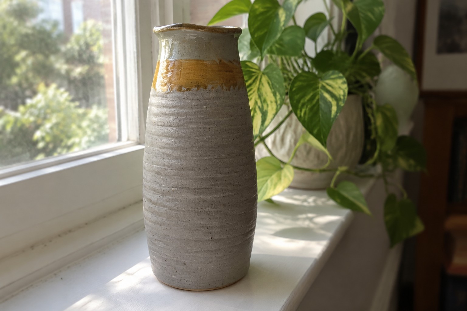 Tall ribbed gray vase with brown glaze on sunny windowsill beside plant.