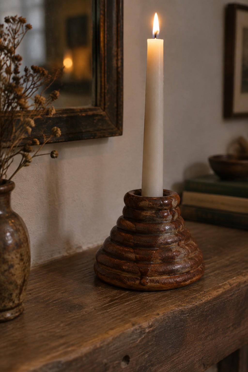 Lit white candle in brown stacked ceramic holder on rustic wooden table with dried flowers