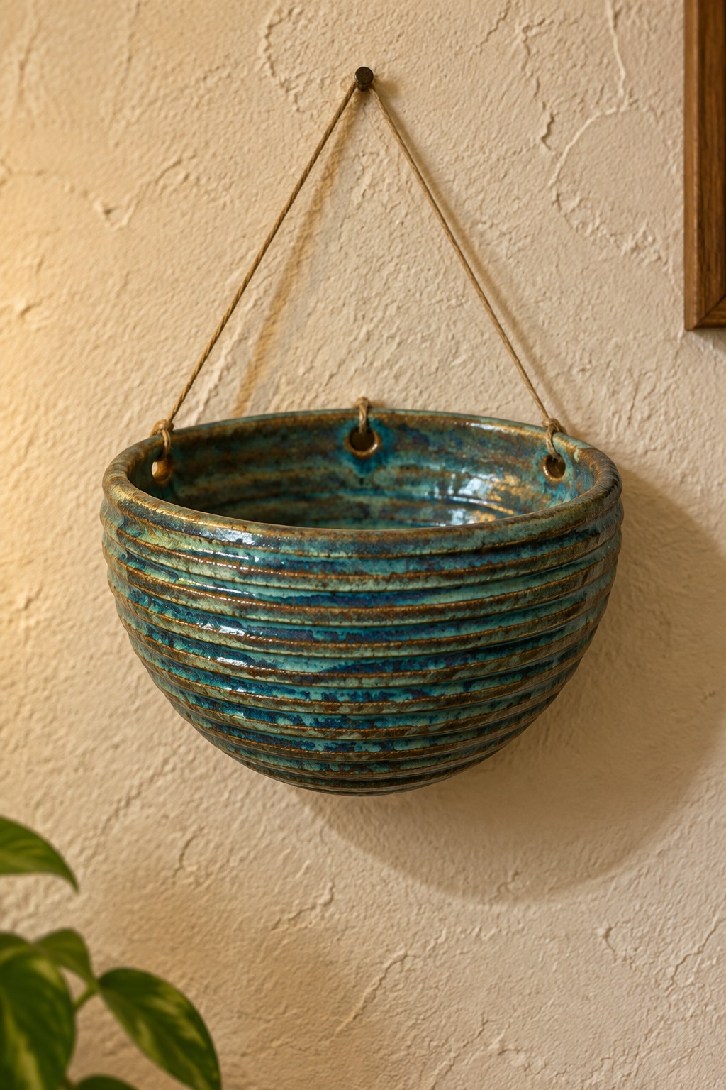 Turquoise ribbed ceramic hanging bowl with brown streaks suspended by ropes on wall.