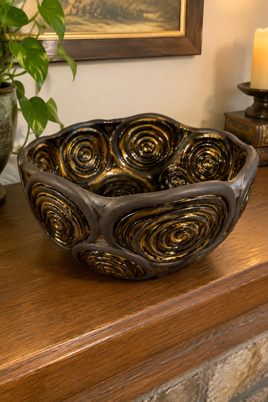 Dark ceramic bowl with golden spiral patterns on wooden surface near plant and candle