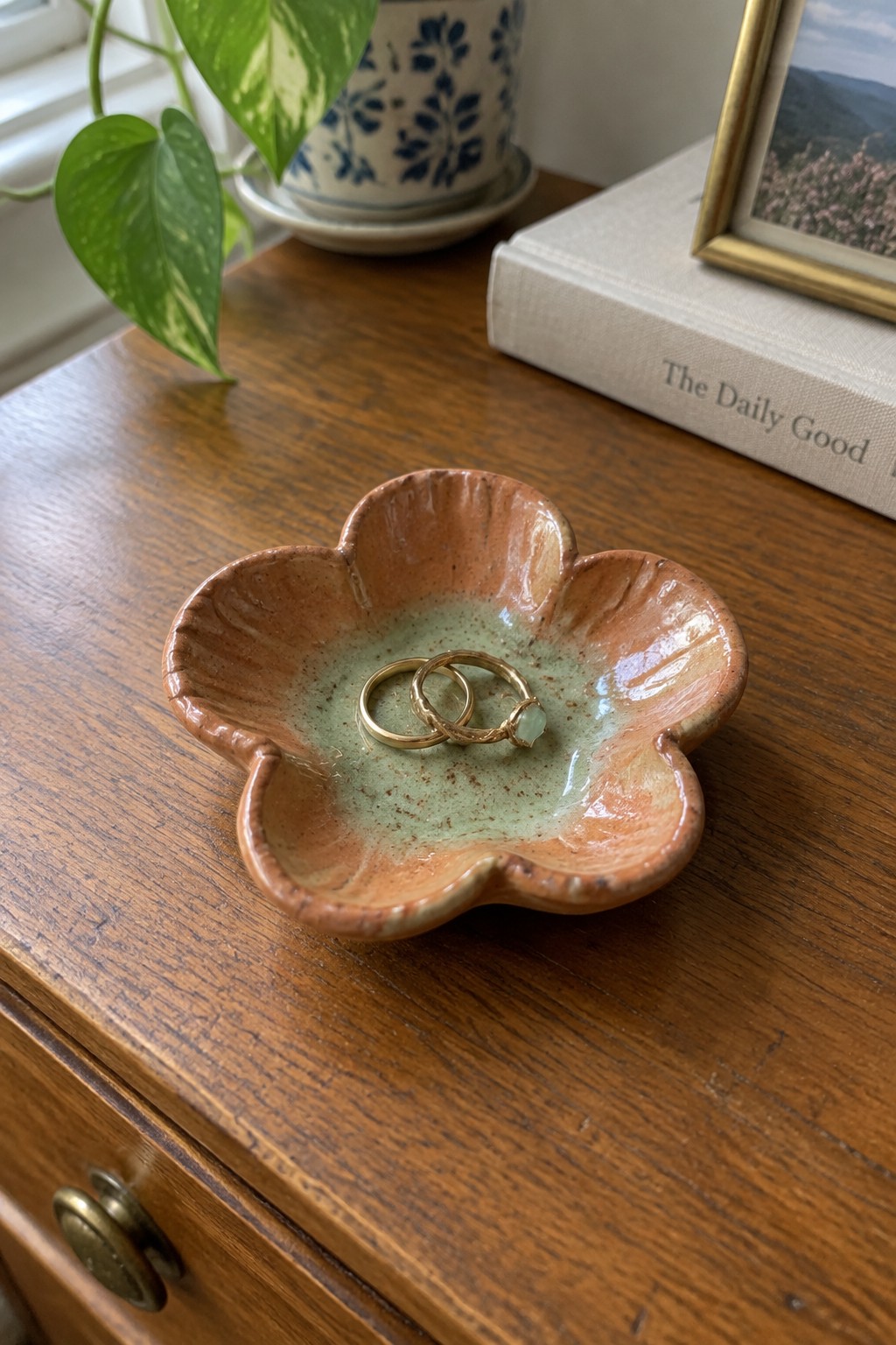 Flower-shaped orange ceramic dish holding gold rings on wooden table with plant
