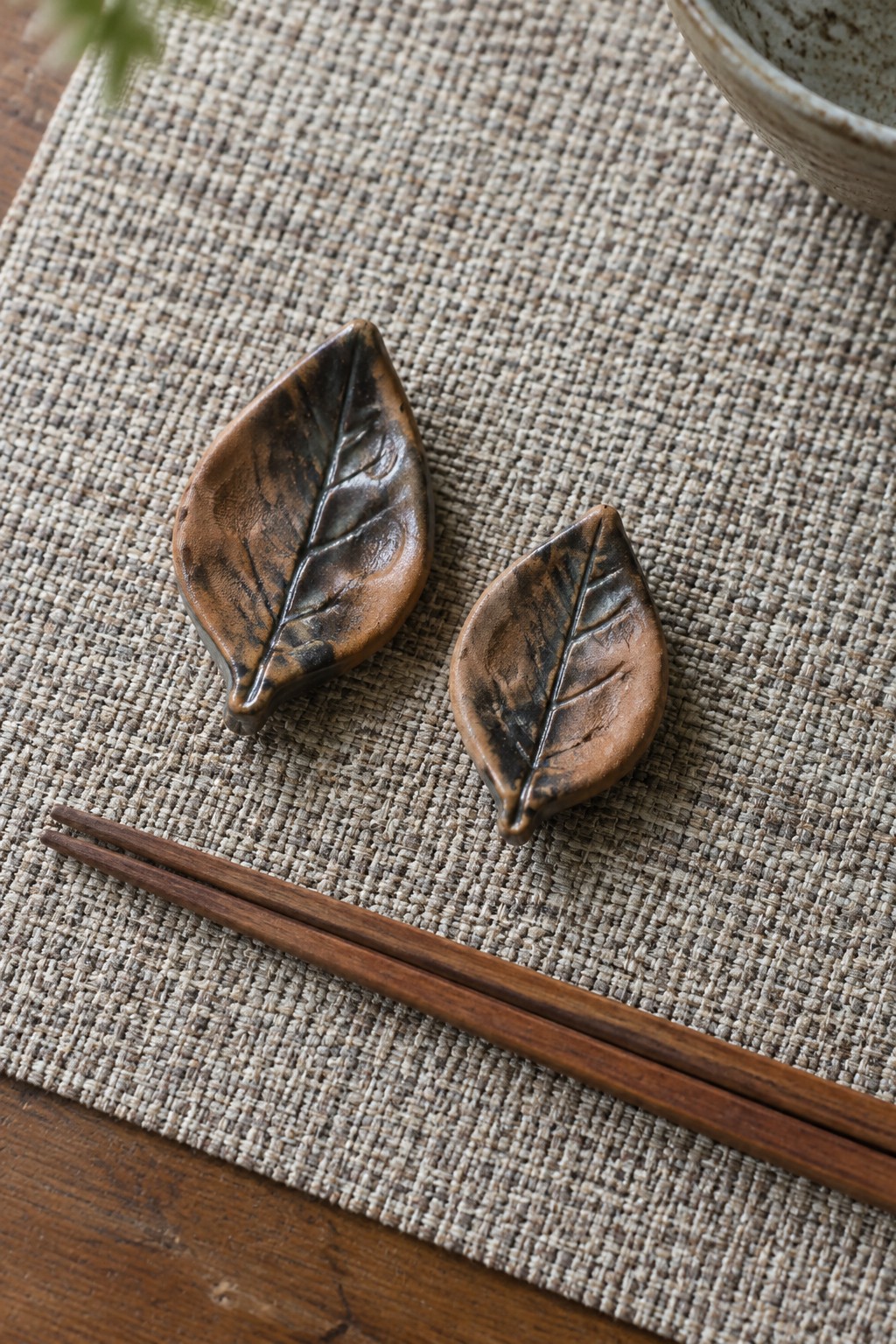 Two brown ceramic leaf chopstick rests with wooden chopsticks on woven placemat
