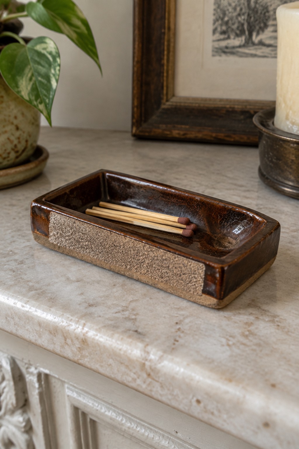 Brown ceramic tray with wooden matches on marble mantel beside plant and candle.
