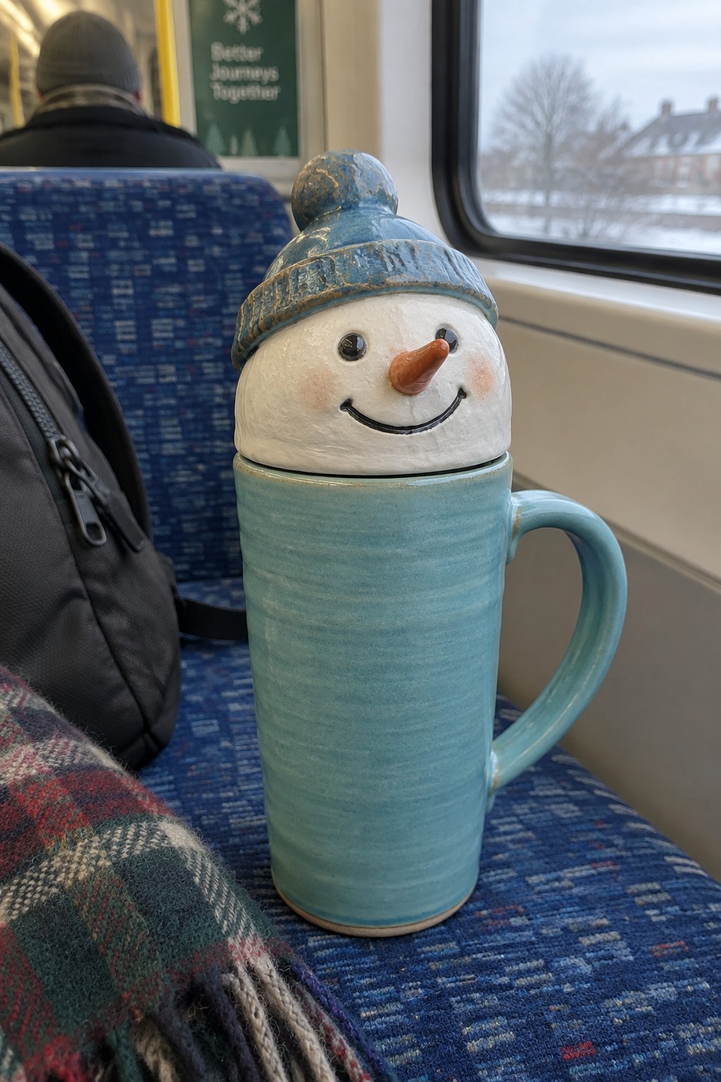 Snowman ceramic mug on blue train seat beside backpack and plaid scarf