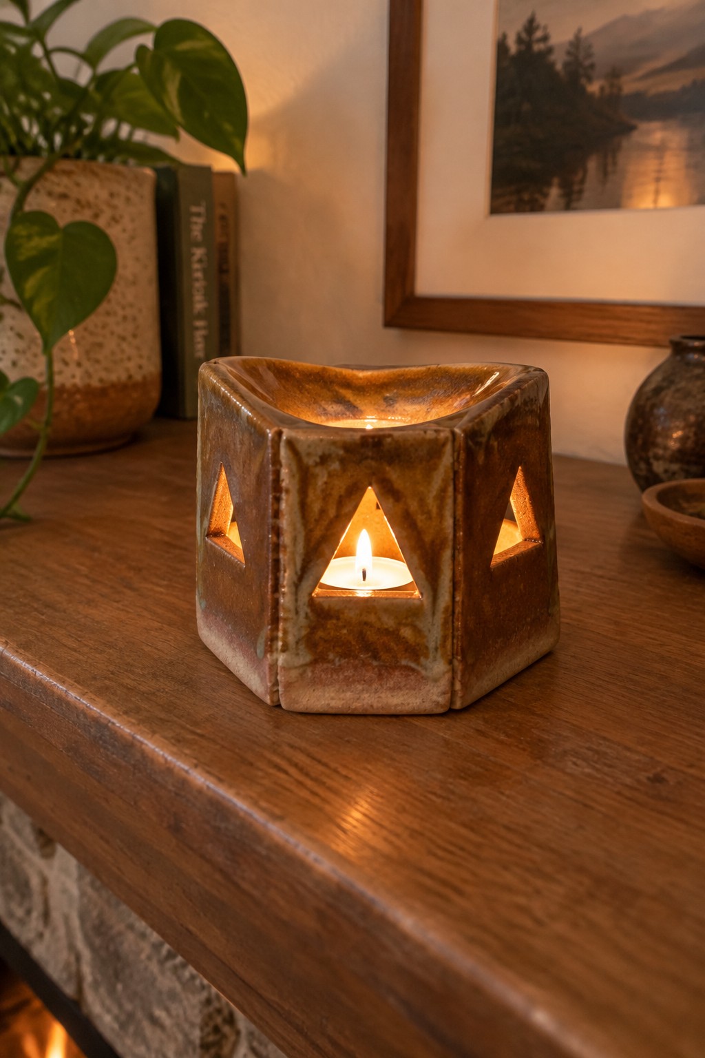 Lit candle in brown ceramic holder with triangular cutouts on wooden table beside plant