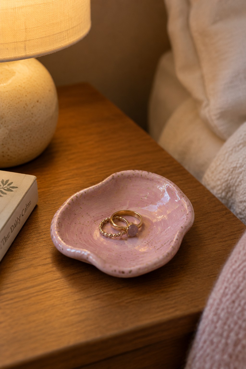 Pink speckled dish holding gold rings on wooden nightstand near lamp and book
