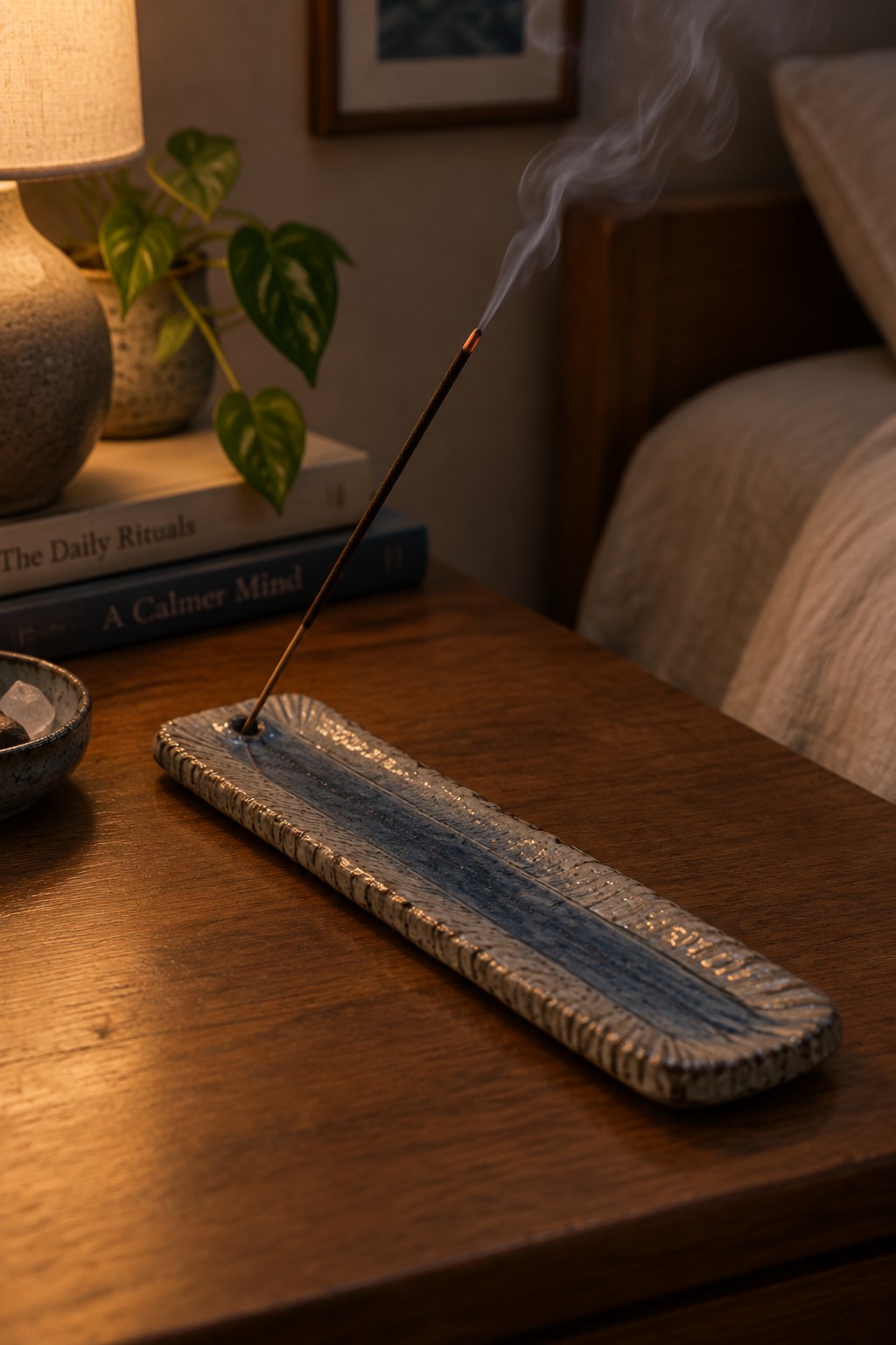 Burning incense stick in blue ceramic holder on wooden nightstand with books and plant