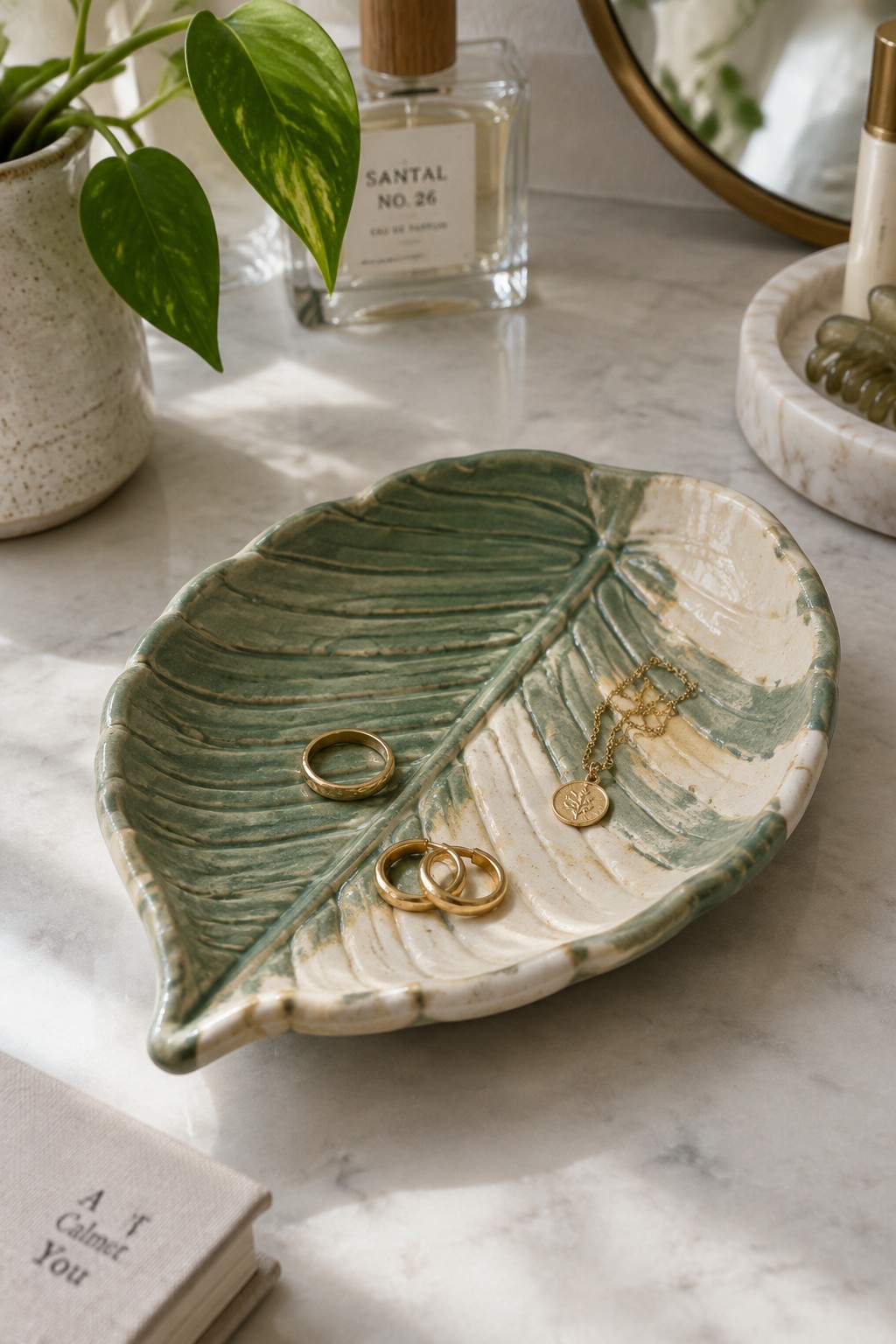 Green leaf-shaped ceramic dish holding gold rings, earrings and necklace on marble