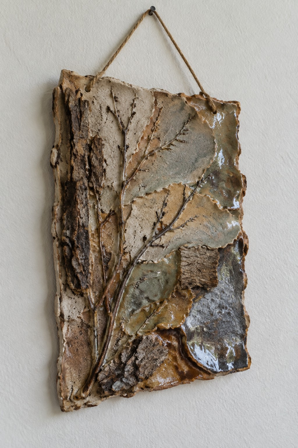 Textured ceramic wall art with raised brown branches on beige and gray glaze, hung by rope.