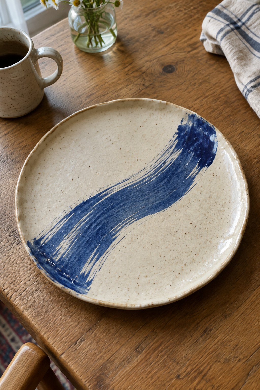 Beige plate with blue brushstroke beside coffee mug and daisy vase on wooden table
