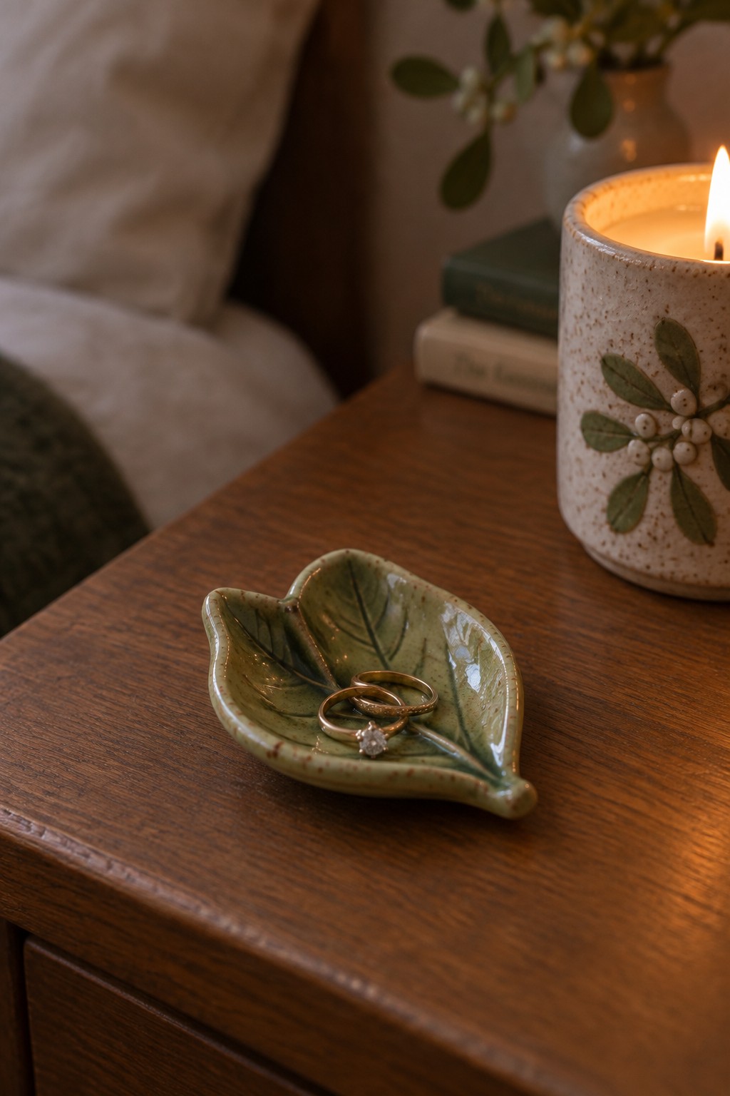 Gold rings rest in a green leaf-shaped ceramic dish beside a lit candle on wood.