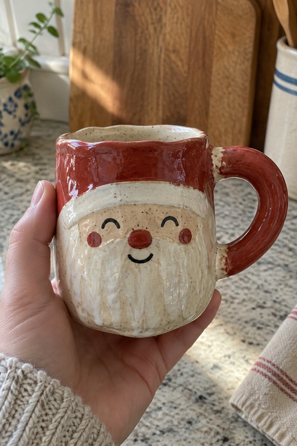 Hand in beige knit sweater holding red-and-white Santa Claus ceramic mug