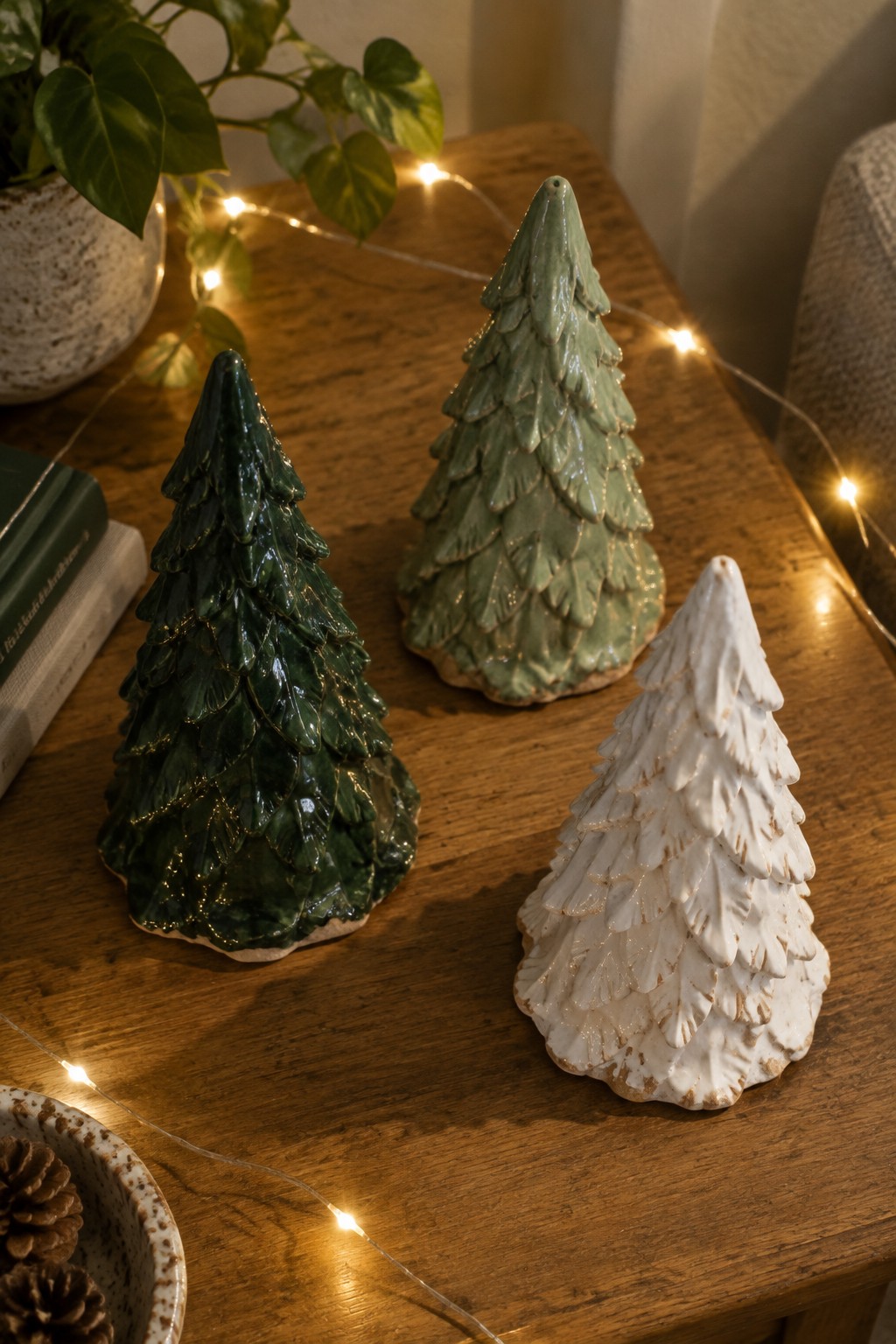 Three ceramic Christmas trees in dark green, sage, and white on wooden table with lights.
