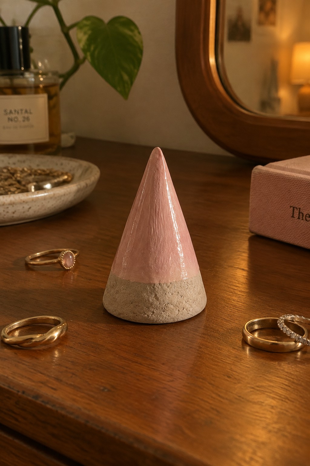 Pink cone sculpture with gray base on wooden table amid gold rings and jewelry.
