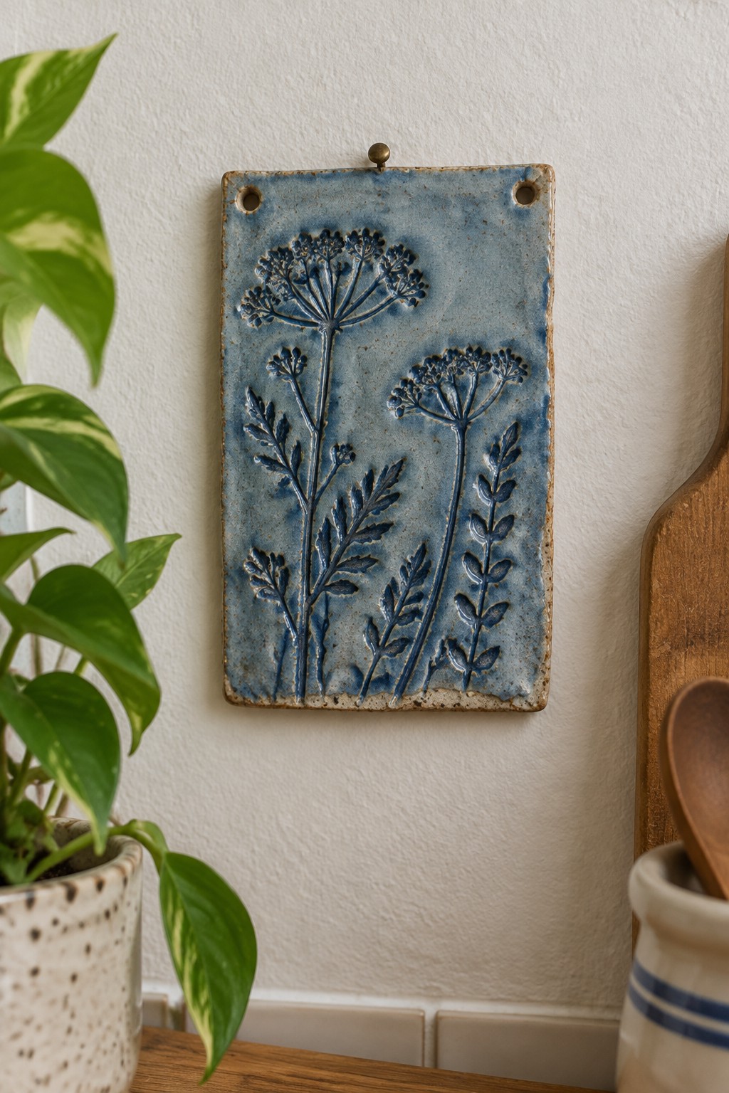 Blue ceramic tile with embossed flowers hanging on wall beside potted plant