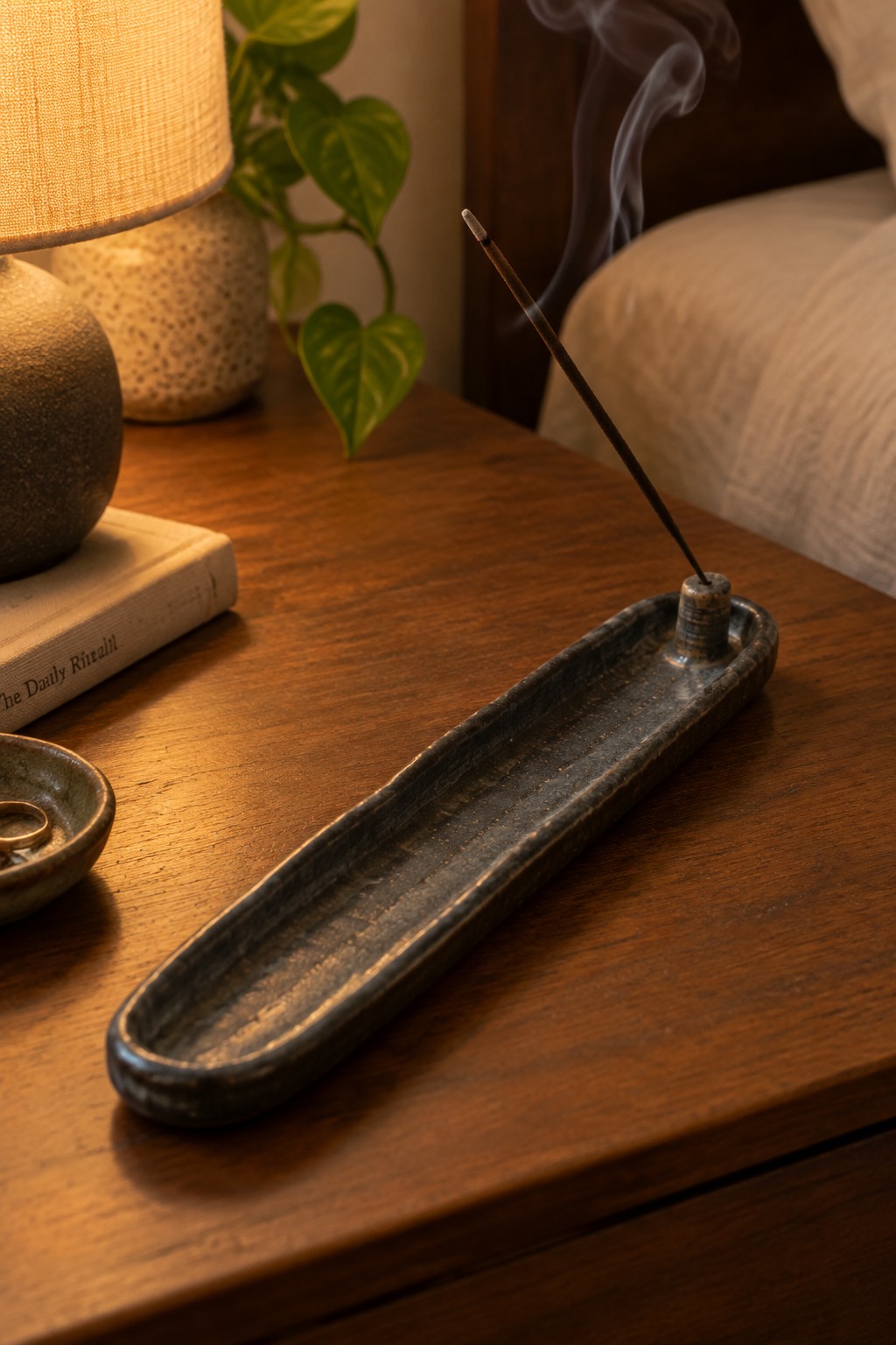 Burning incense in long dark ceramic holder on wooden bedside table with lamp and book