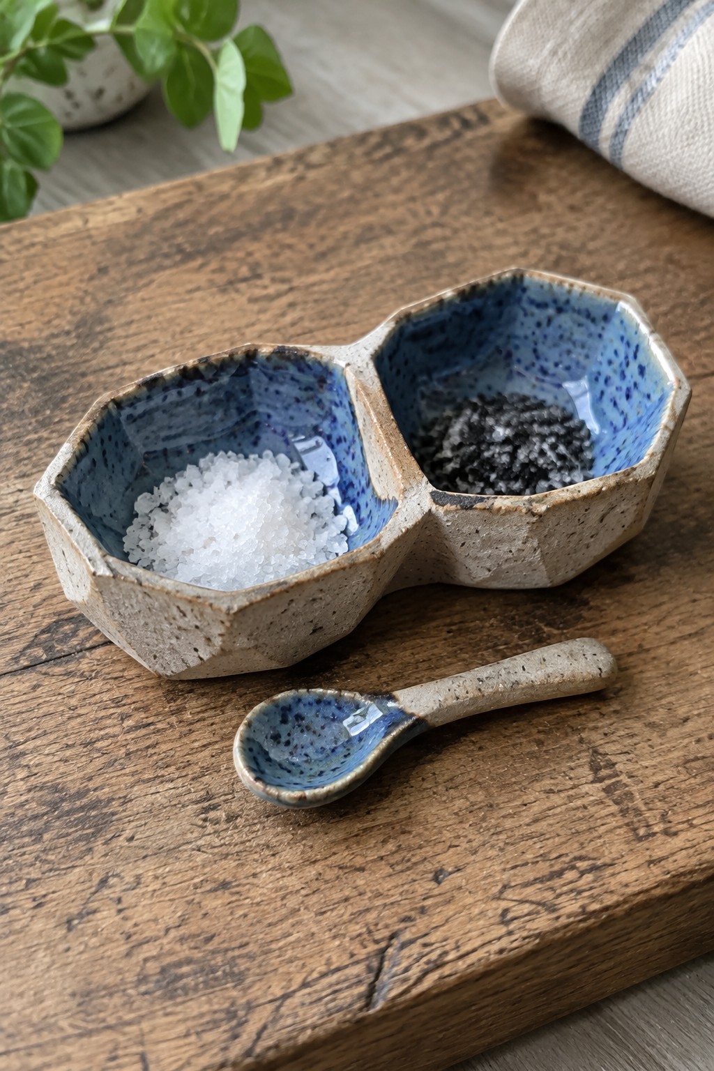 Blue speckled ceramic double bowl with white and black salts beside matching spoon on wood.