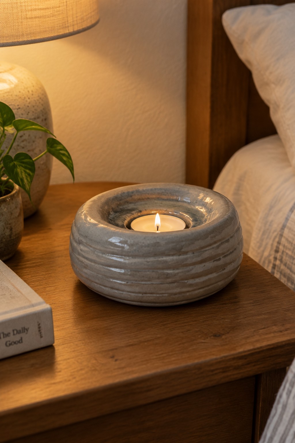 Lit candle in ribbed gray ceramic holder on wooden nightstand with plant and book