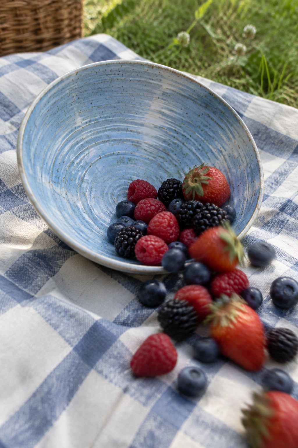 Blue speckled bowl of mixed berries on checkered picnic blanket with scattered fruit