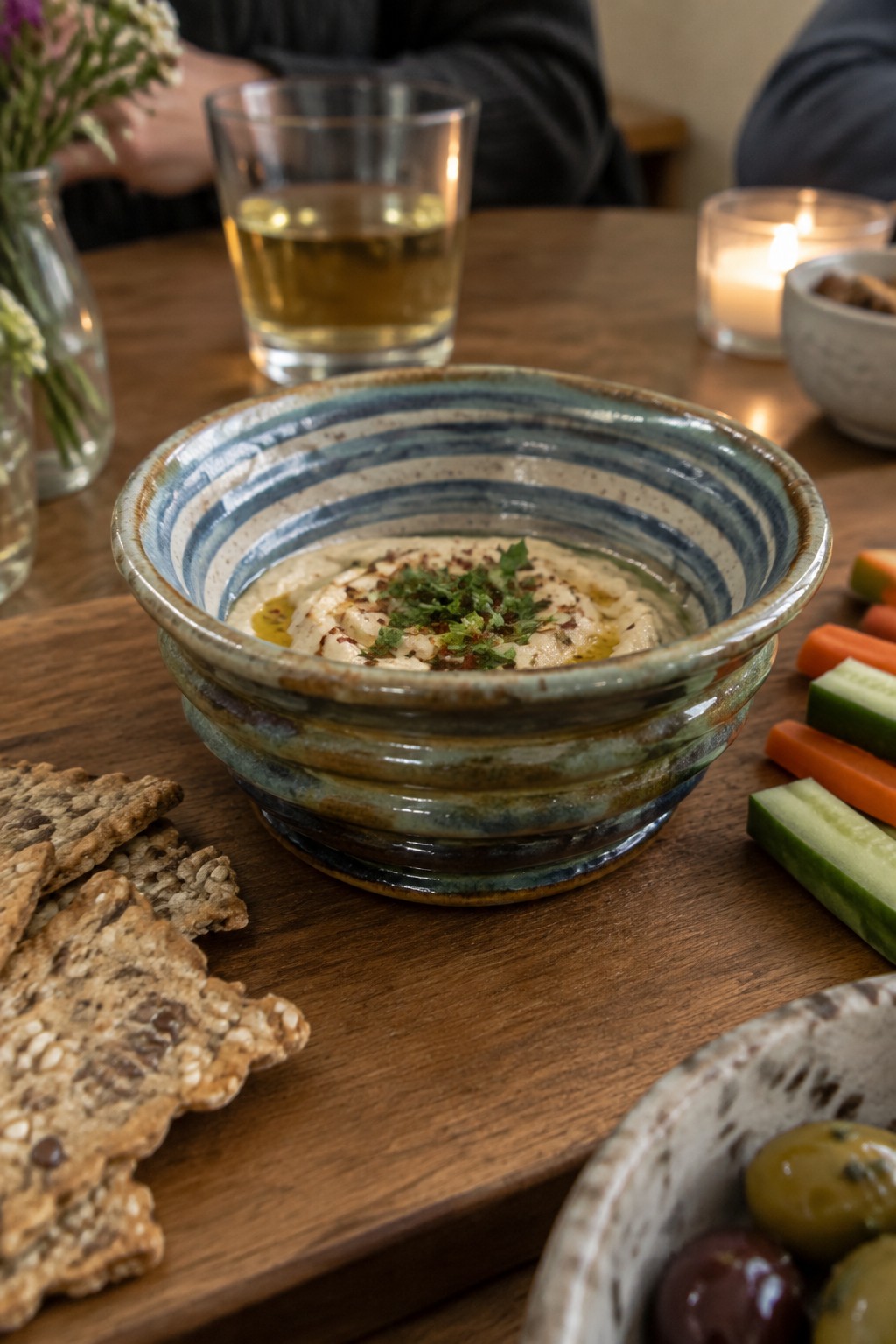 Striped ceramic bowl of hummus with crackers, olives, and vegetable sticks