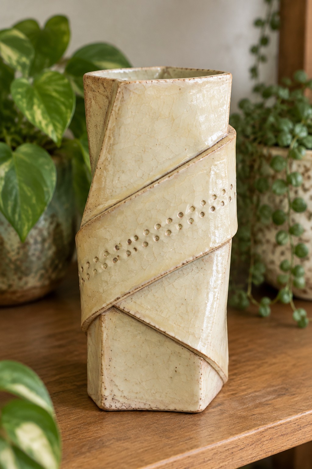 Creamy twisted ceramic vase with dotted bands on wooden table amid plants.