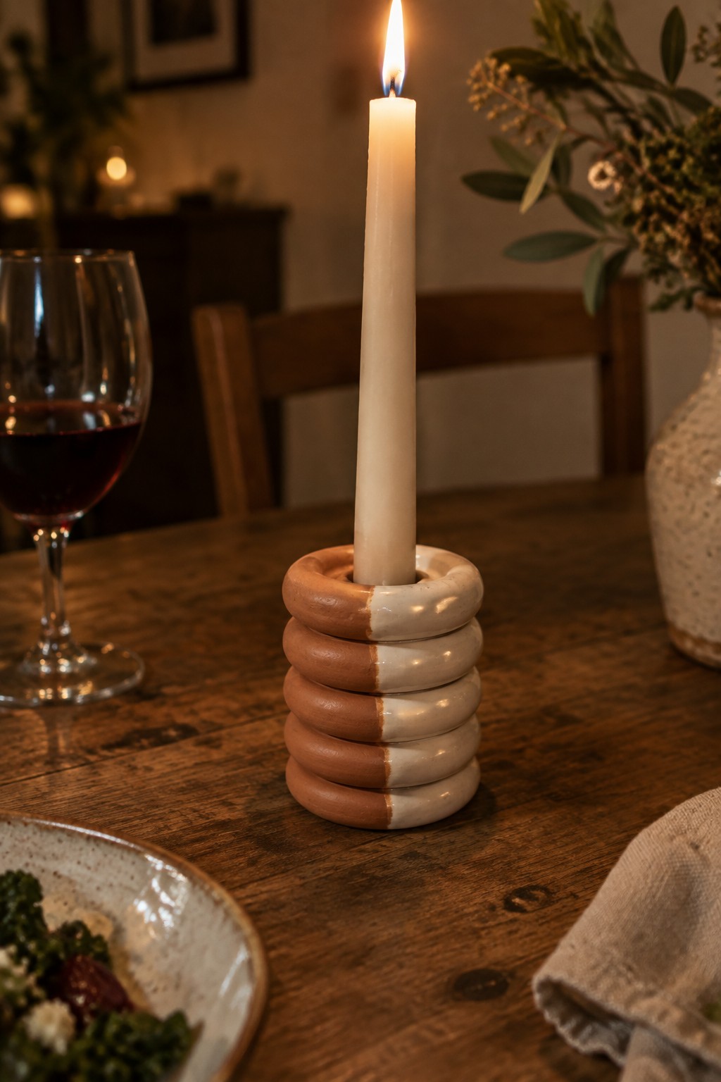 Lit candle in stacked brown-and-white ceramic holder on rustic wooden table with wine glass