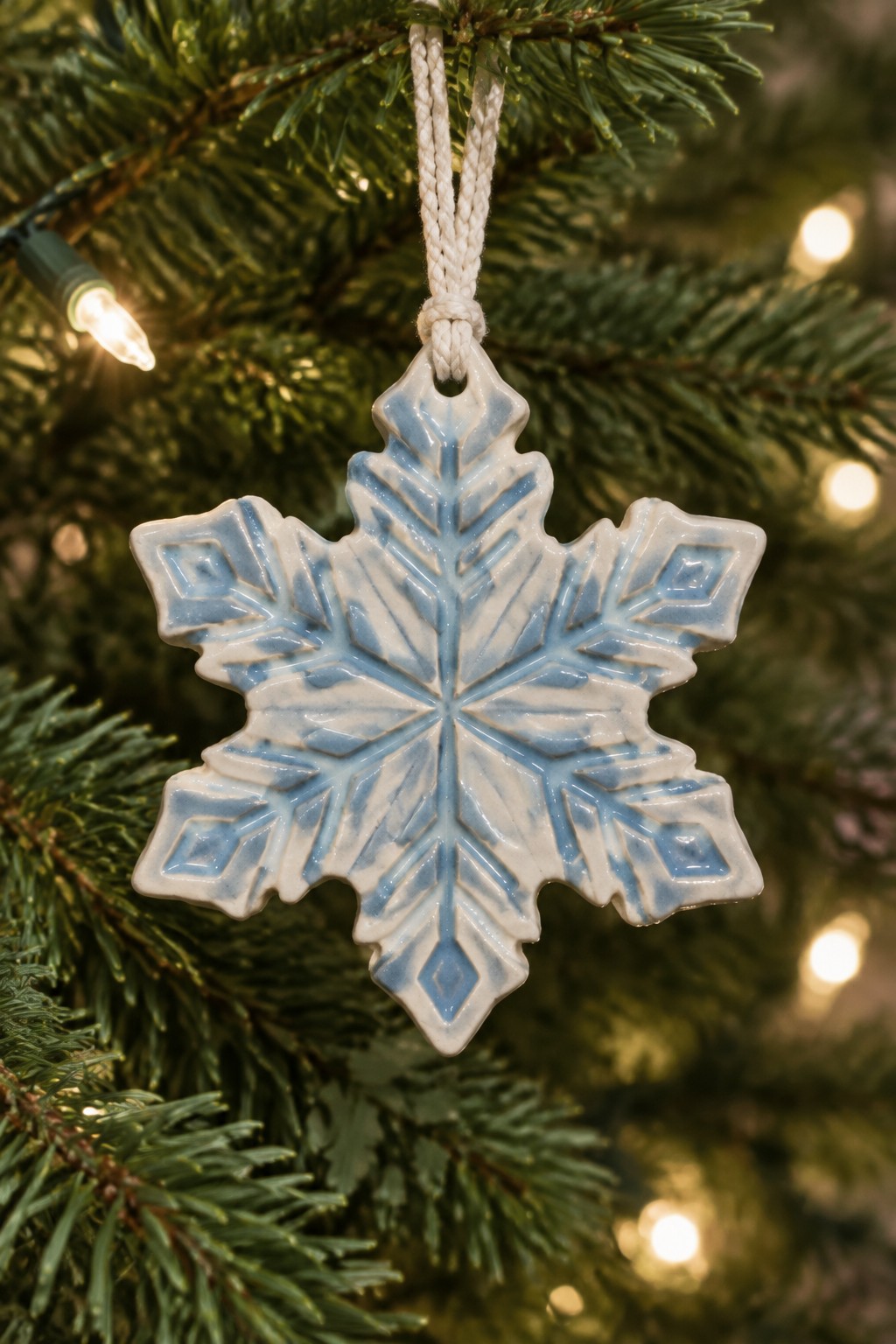 Ceramic snowflake ornament in blue and white hanging from lit Christmas tree branch.