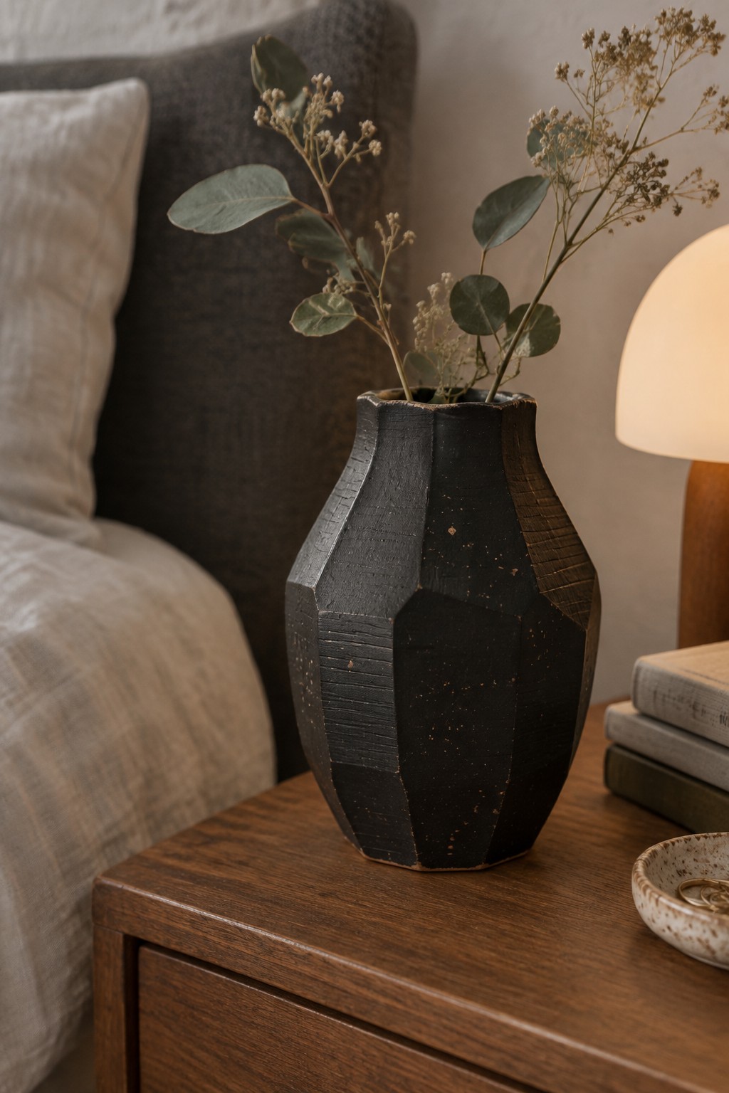 Black faceted vase with dried eucalyptus on wooden nightstand beside bed