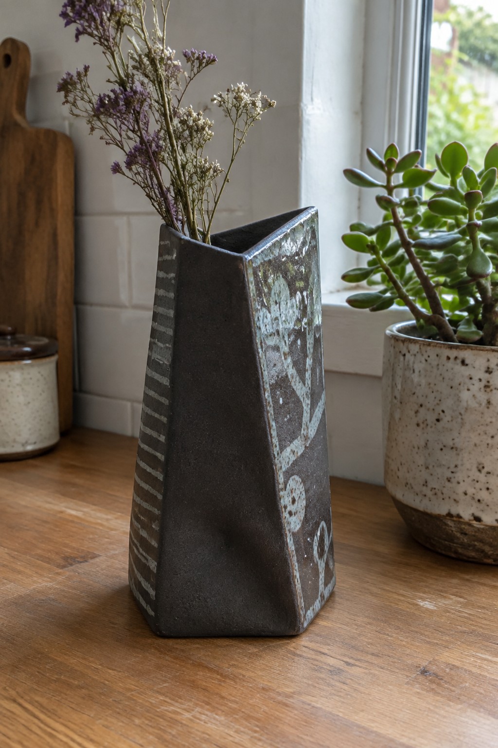 Geometric dark ceramic vase with dried purple flowers on wooden counter near succulent