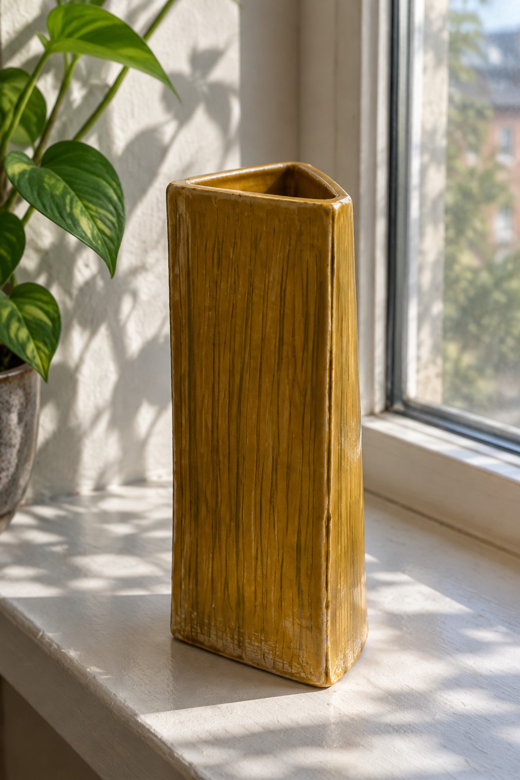 Tall mustard-yellow textured ceramic vase on sunlit windowsill beside green plant