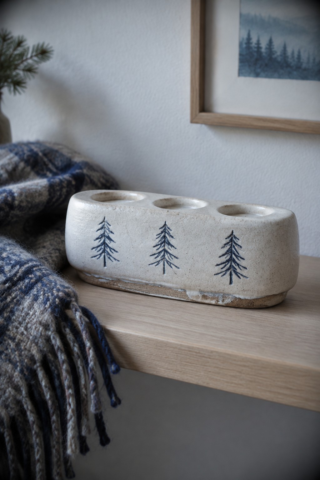 A handmade ceramic candle holder with three holes and three blue pine tree designs rests on a wooden surface.