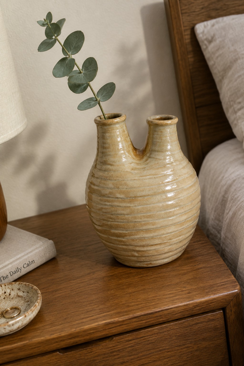 Ribbed beige double-neck ceramic vase with eucalyptus on wooden nightstand beside book and dish