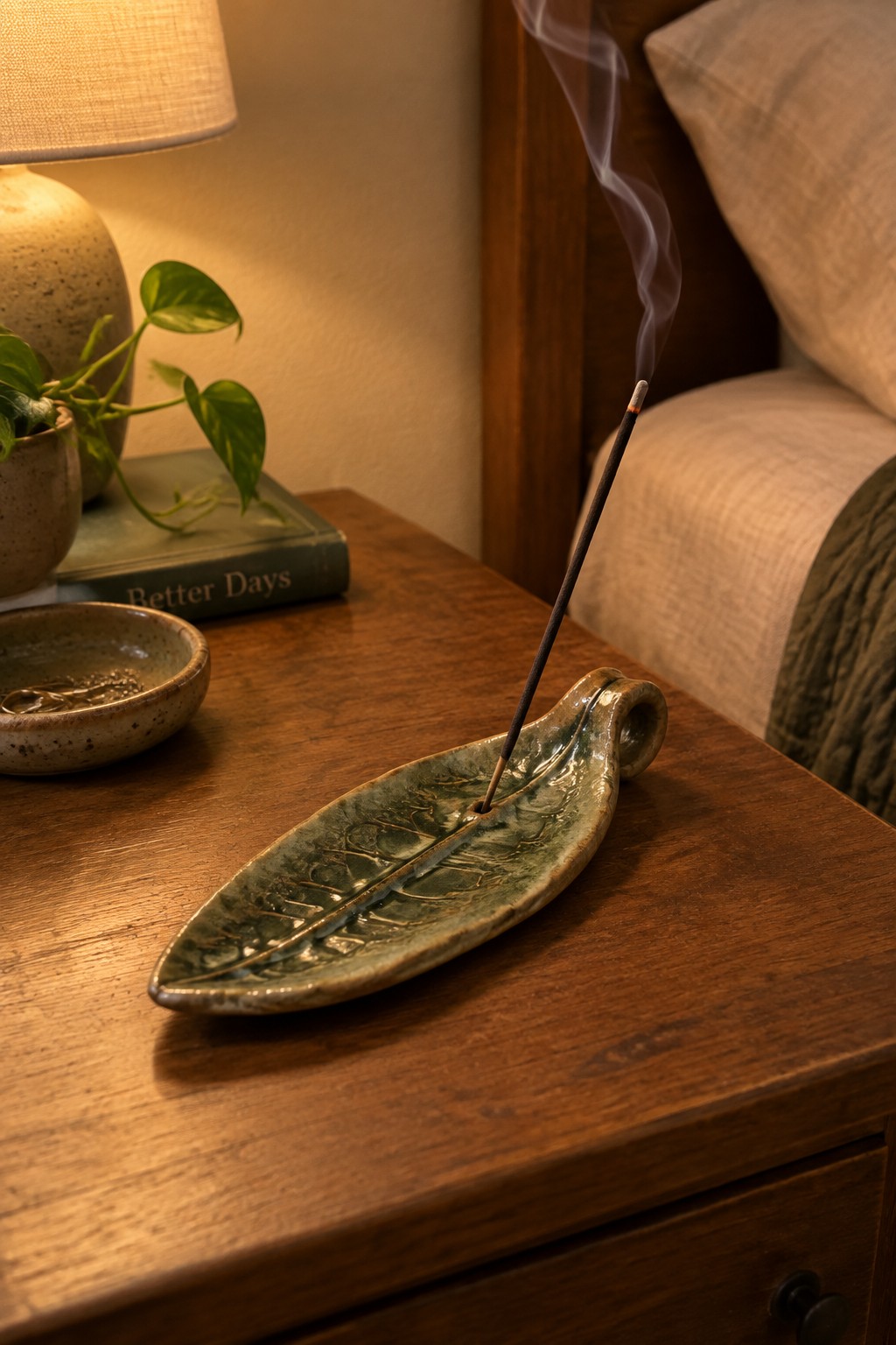 Green ceramic leaf incense holder with burning stick and rising smoke on wooden nightstand