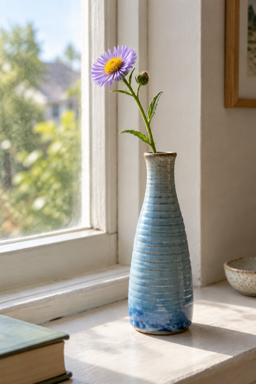Blue ribbed vase with purple flower on sunlit windowsill beside green book